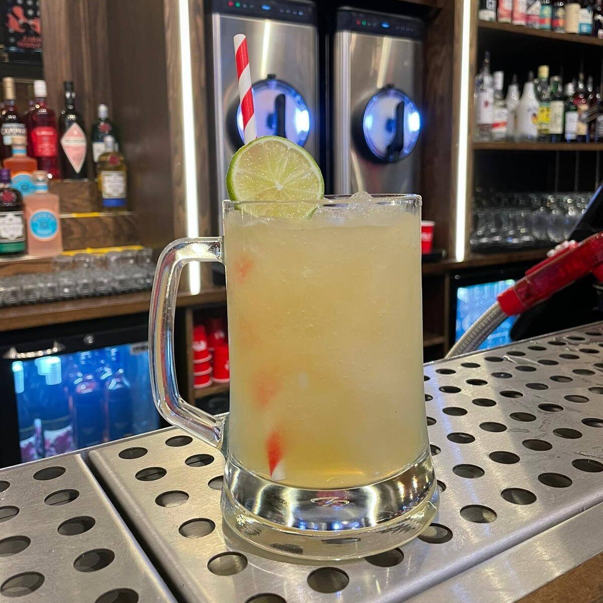 A frosty cocktail in a glass mug, garnished with a lime slice and a red-and-white straw, sits on a bar counter with liquor bottles behind.