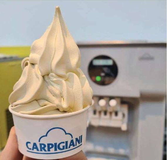 A hand holds a cup of creamy, swirled soft-serve gelato in a Carpigiani-branded container, with a gelato machine in the background.