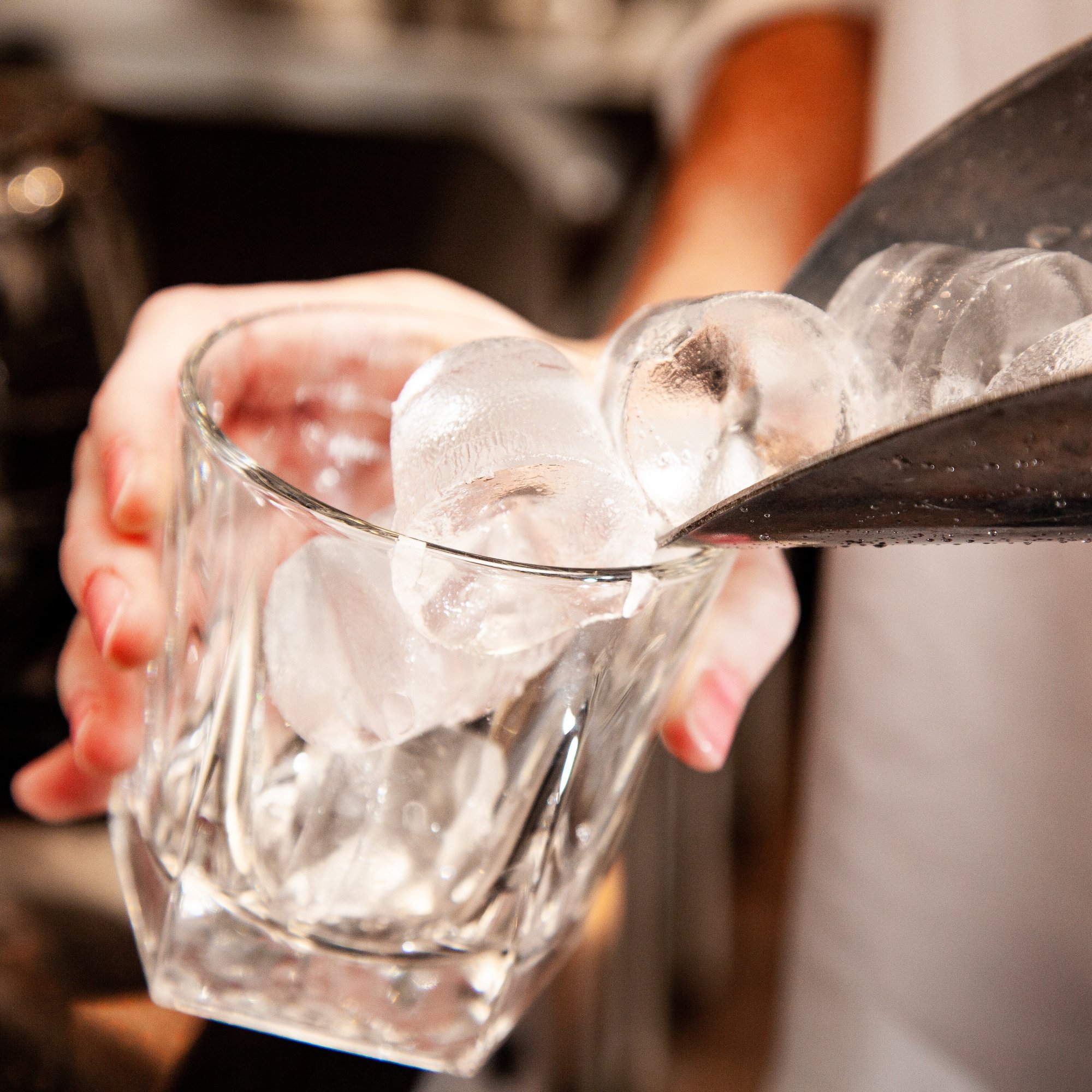 A bartender or server pours round ice cubes from a metal scoop into a clear glass, with a blurred background of a bar or kitchen setting.