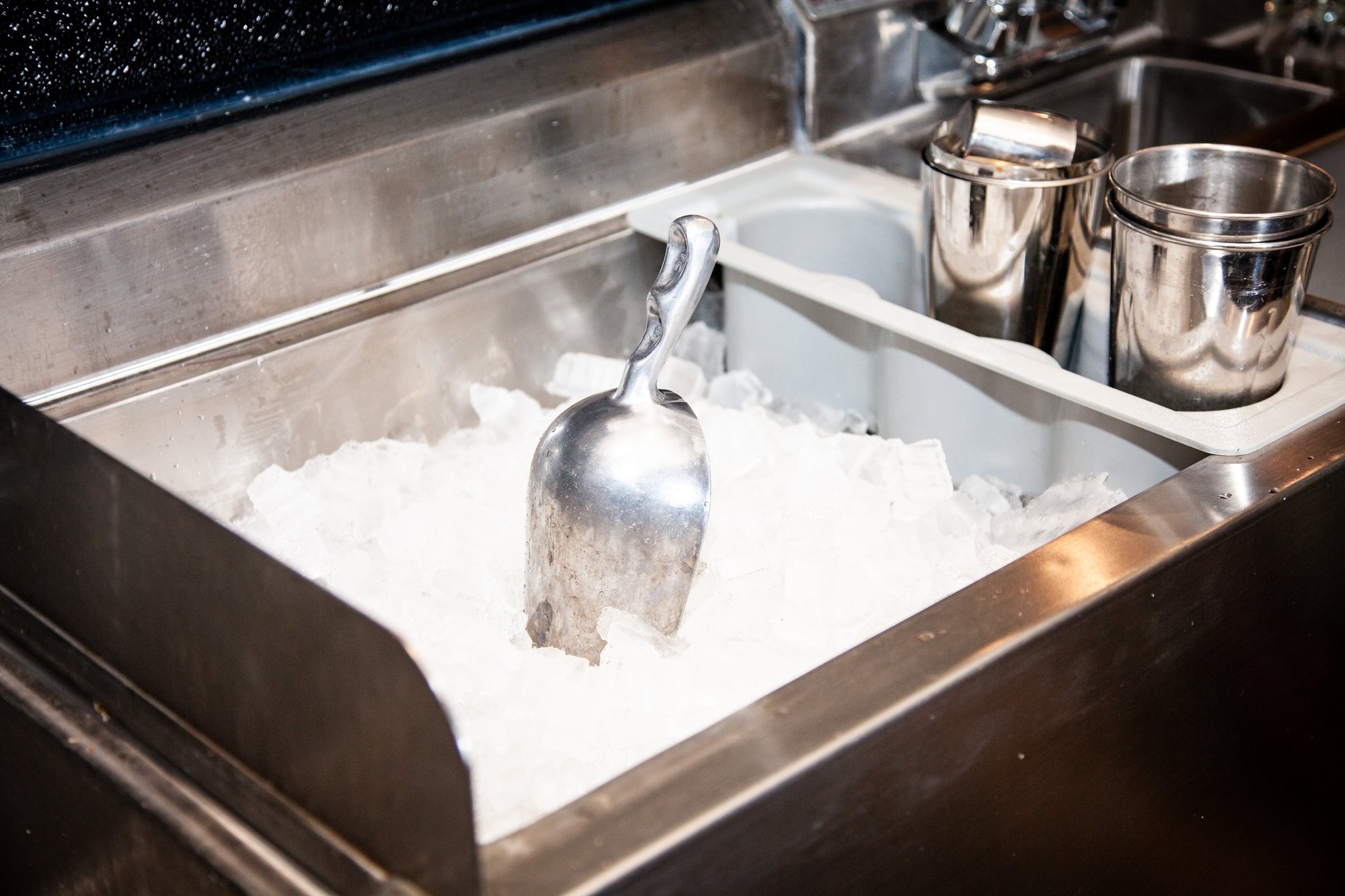 A stainless steel ice bin filled with crushed ice, featuring a metal scoop resting on top, with metal cups and a sink in the background.