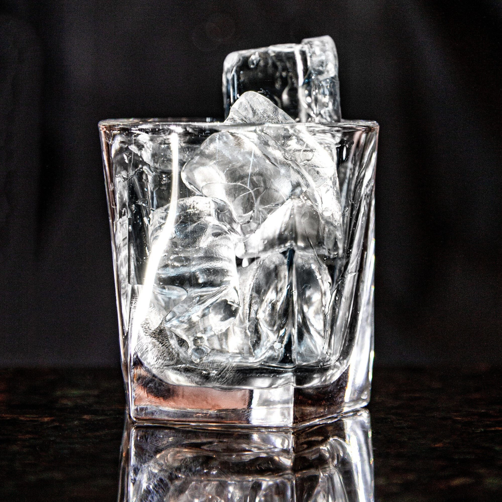 A clear glass filled with irregularly shaped ice cubes sits on a reflective surface against a dark background.