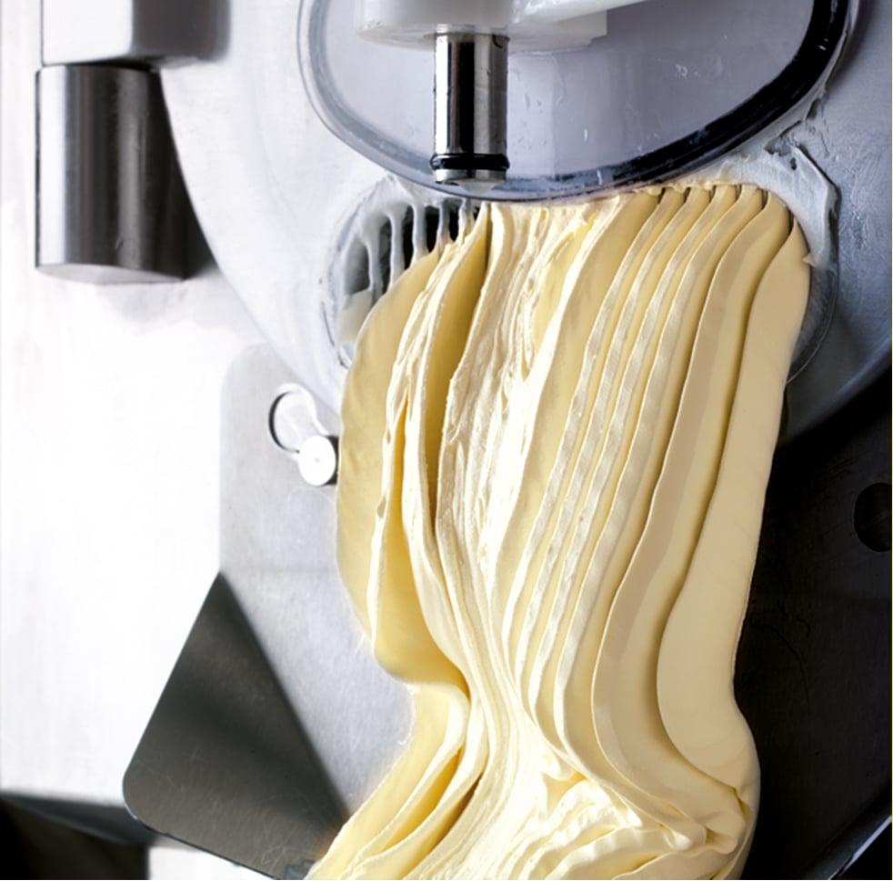 Freshly churned gelato with smooth, delicate folds emerges from a commercial ice cream machine, creating a soft, creamy texture.