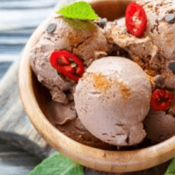 A wooden bowl holds scoops of chocolate ice cream topped with red chili slices, cinnamon, chocolate chips, and a fresh mint leaf.