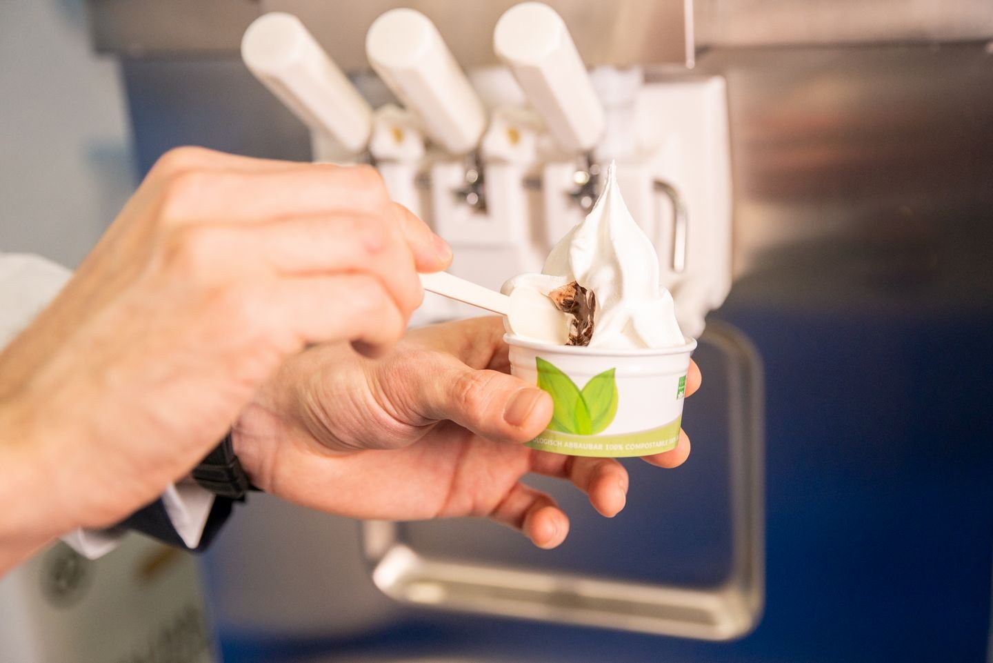 A person holds a small cup of swirled soft-serve gelato with a wooden spoon, in front of a commercial soft-serve machine.