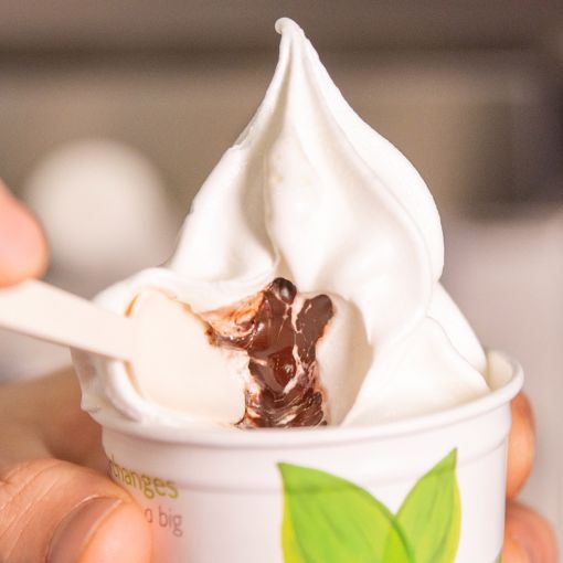 A close-up of creamy white soft-serve gelato in a cup, revealing a rich chocolate filling as a wooden spoon scoops into it.