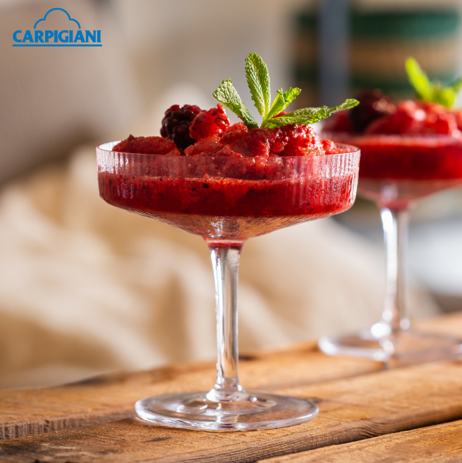 A coupe glass filled with a red berry granita, garnished with fresh mint and blackberries, sits on a rustic wooden surface.